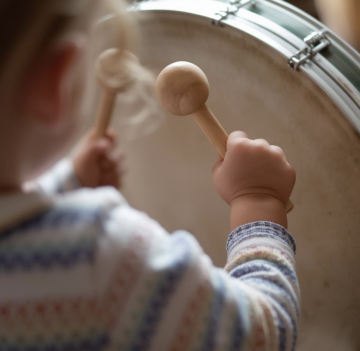 Ein Kleinkind das von hinten sichtbar ist und auf einer Rahmentrommel mit Schlägeln trommelt(1)